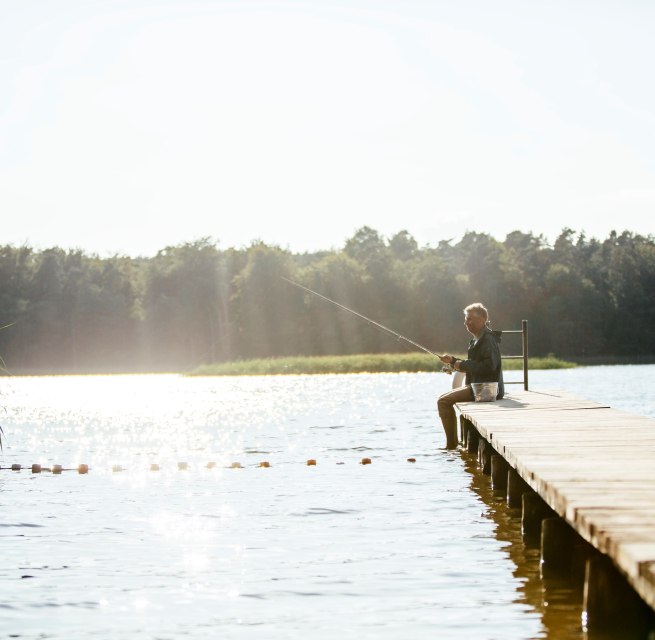 A man sits on a wooden jetty on a calm lake and fishes while the sun makes the water glisten. // Relaxed hours in nature - An angler enjoys the tranquillity of the lake on a wooden jetty, surrounded by glistening water and a dense forest backdrop. // © MV-T/Roth A man sits on a wooden jetty on a calm lake and fishes while the sun makes the water glisten.