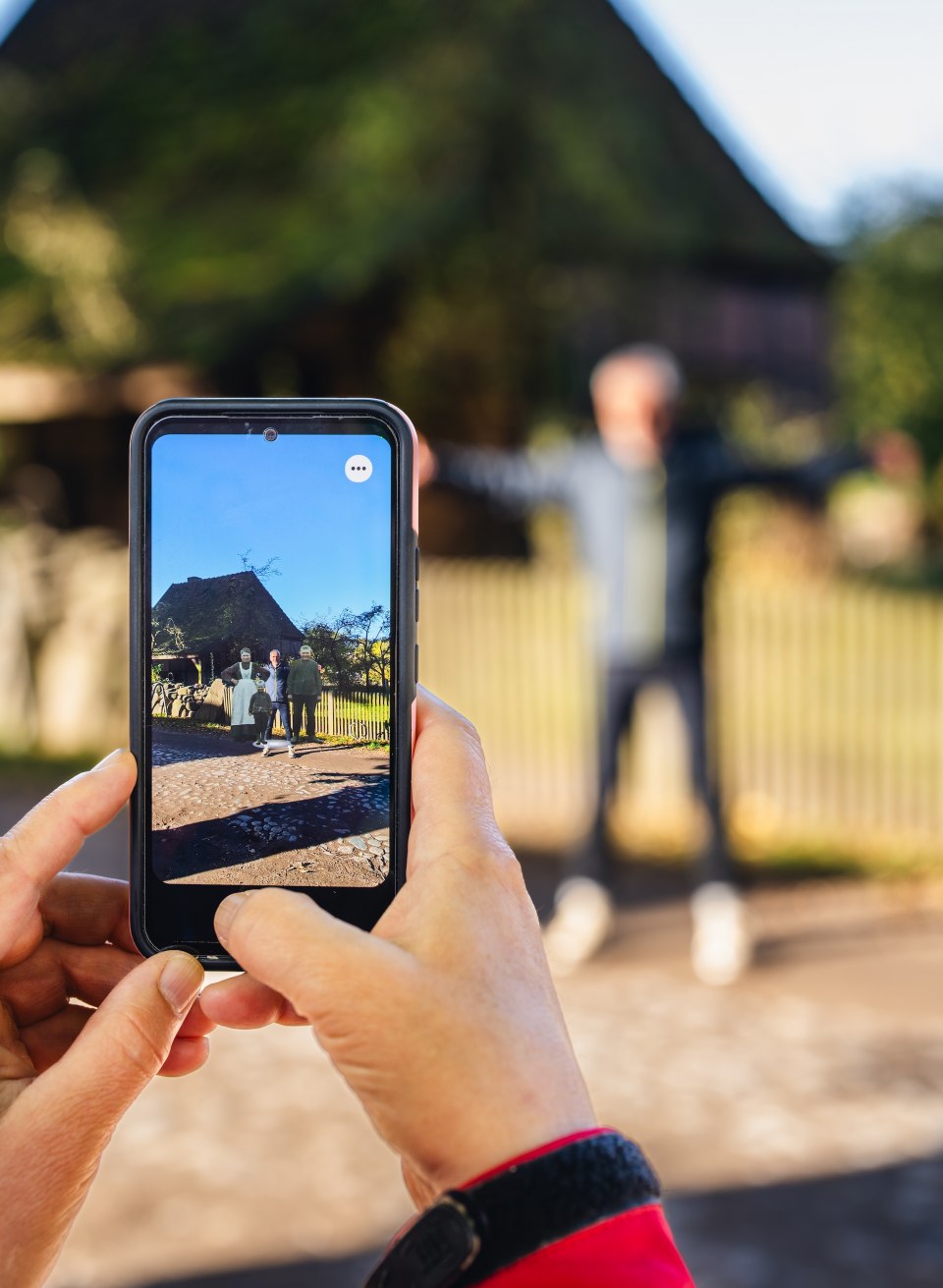 Een persoon gebruikt een smartphone om een historisch gebouw in het openluchtmuseum Schwerin-Mue&szlig; te fotograferen, terwijl een andere persoon op de achtergrond poseert.