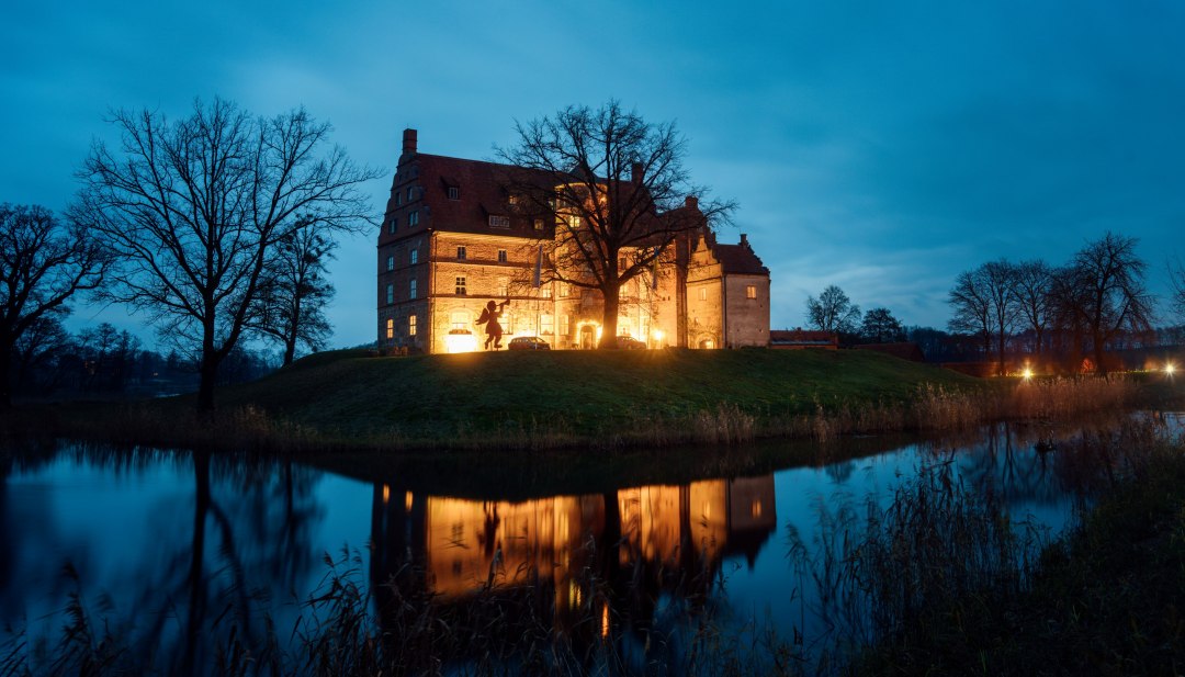 Atmospherically illuminated Ulrichshusen Castle at Christmas time, surrounded by trees and a moat with reflections in the evening light.