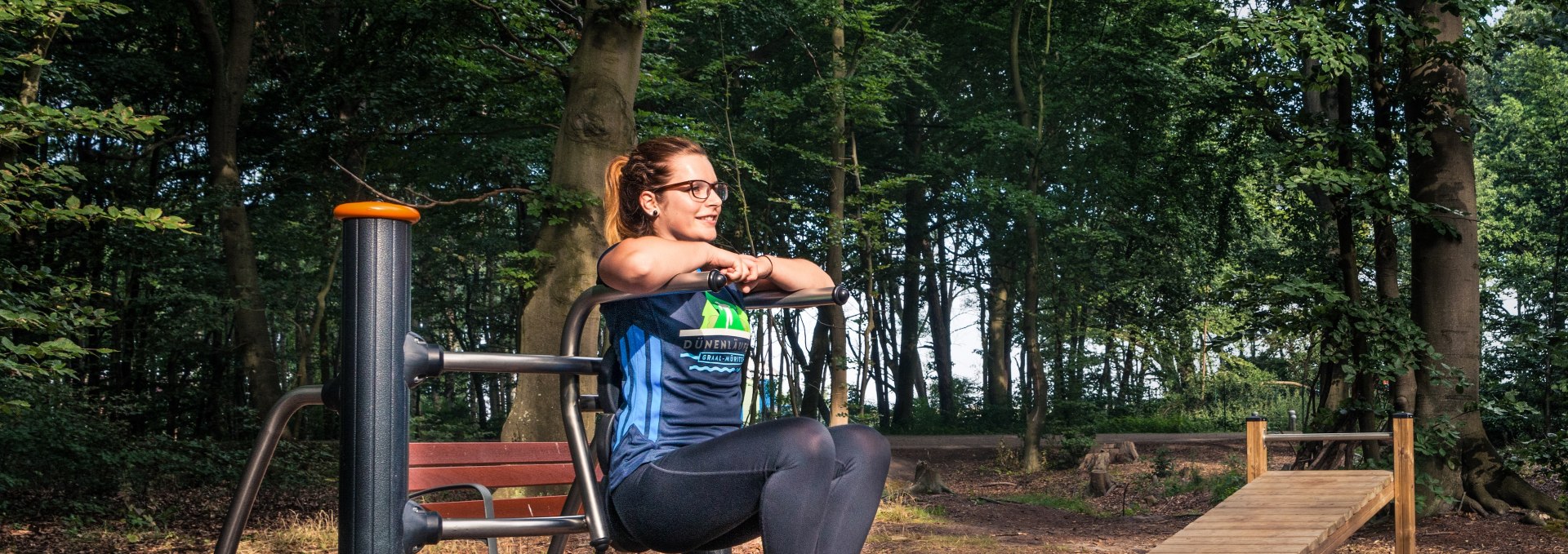 Kobieta ćwiczy na maszynie fitness w aktywnym lesie Graal-Müritz, otoczona wysokimi drzewami i naturalną leśną atmosferą.