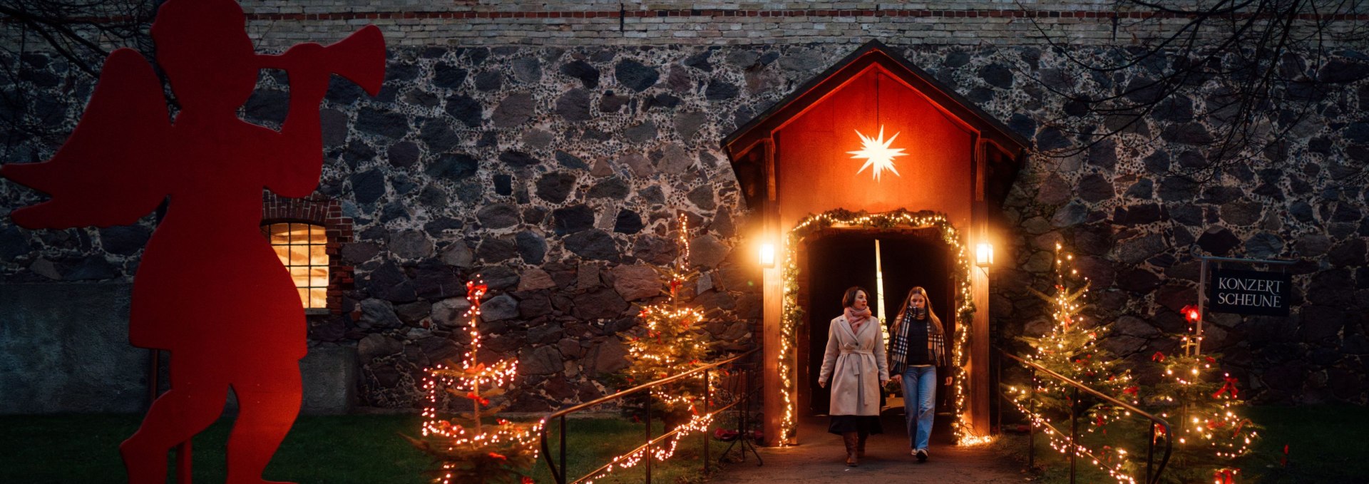 Two women walk out of the Christmas-lit concert barn at Ulrichshusen Castle, flanked by trees of light and a figure of an angel.