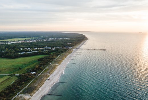 Luchtfoto van de pier en het strand van Graal-Müritz, omgeven door bossen en weilanden, met de Baltische Zee in het licht van een sfeervolle zonsondergang.