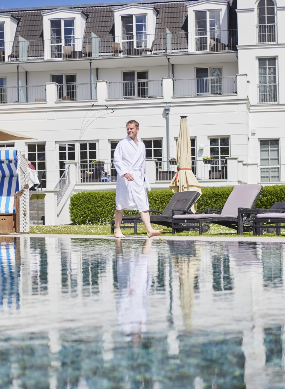 Man in badjas bij het zwembad van Strandhotel Zingst met ligstoelen en strandstoel voor de gevel in zeestijl.