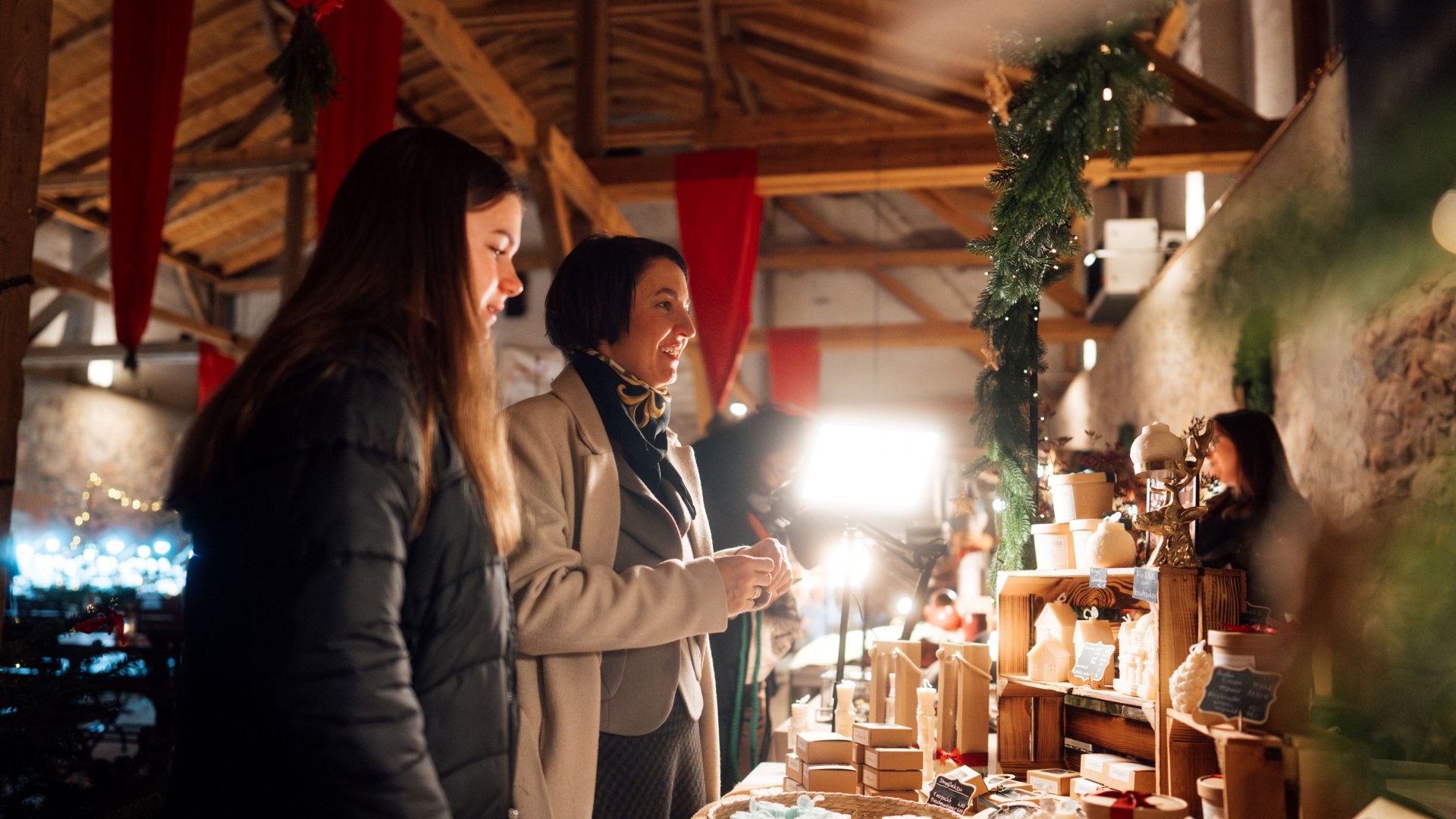 Two women stand at a market stall decorated for Christmas with handmade soaps, candles and gifts and enjoy the festive atmosphere.