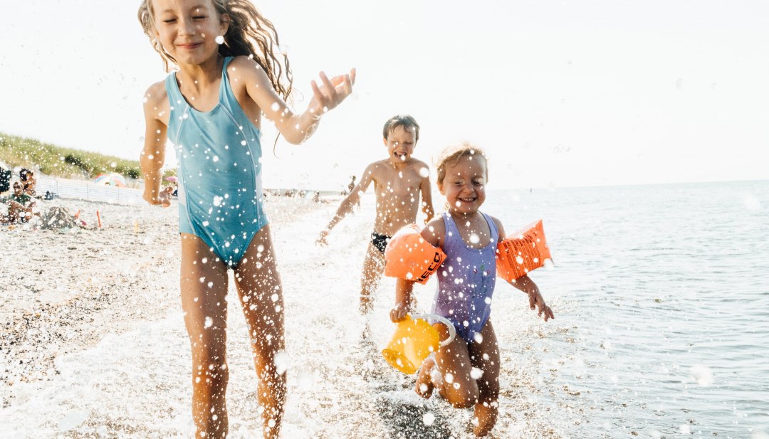 Drie kinderen rennen lachend door het ondiepe water van de Oostzee op het strand van Dierhagen, met opspattend water op de voorgrond. // Kinderen spetteren en rennen door de ondiepe Oostzee in Dierhagen – luchtig, levendig, vol zomerse momenten. // © MV-T/Roth Drie kinderen rennen lachend door het ondiepe water van de Oostzee op het strand van Dierhagen, met opspattend water op de voorgrond.