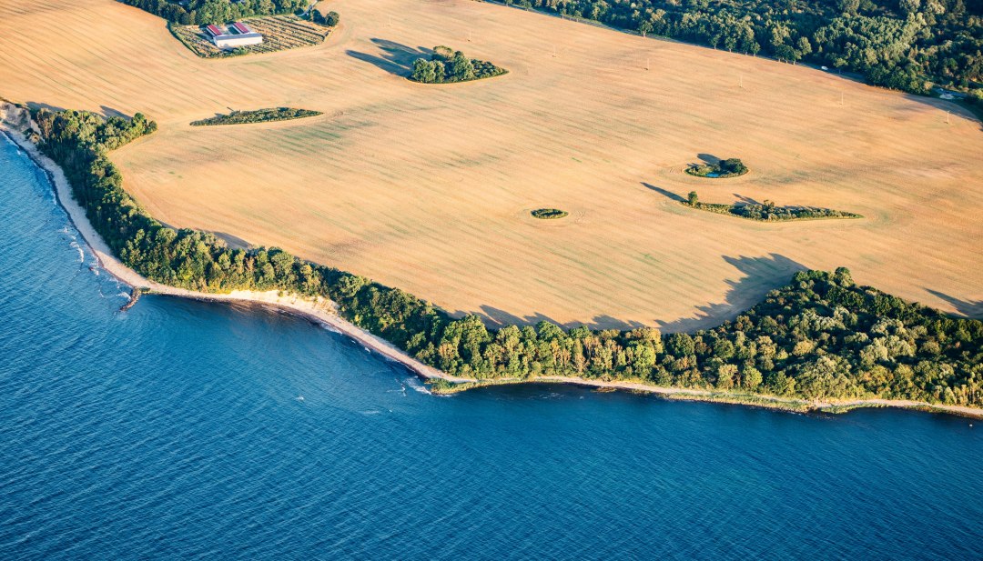 Luchtfoto van de steile kust van Rügen bij Glowe toont het contrast tussen gouden velden en turkooisblauwe zee, omringd door groene bossen. // Majestueuze kliffen bij Glowe op Rügen, waar gouden velden en een azuurblauwe zee samen een onvergetelijk panorama vormen. De uitgestrektheid van de Baltische Zee en de krachtige natuur van Duitslands grootste eiland inspireren tot nieuwe horizonten. // © MV-T/Gänsicke Luchtfoto van de steile kust van Rügen bij Glowe toont het contrast tussen gouden velden en turkooisblauwe zee, omringd door groene bossen.
