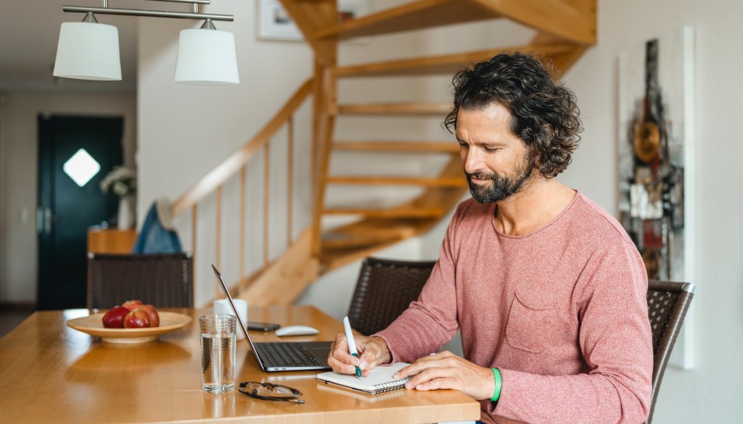 A man sits at a wooden table in a vacation home, working on his laptop and taking notes - ideal conditions for a workation on the island of Poel.