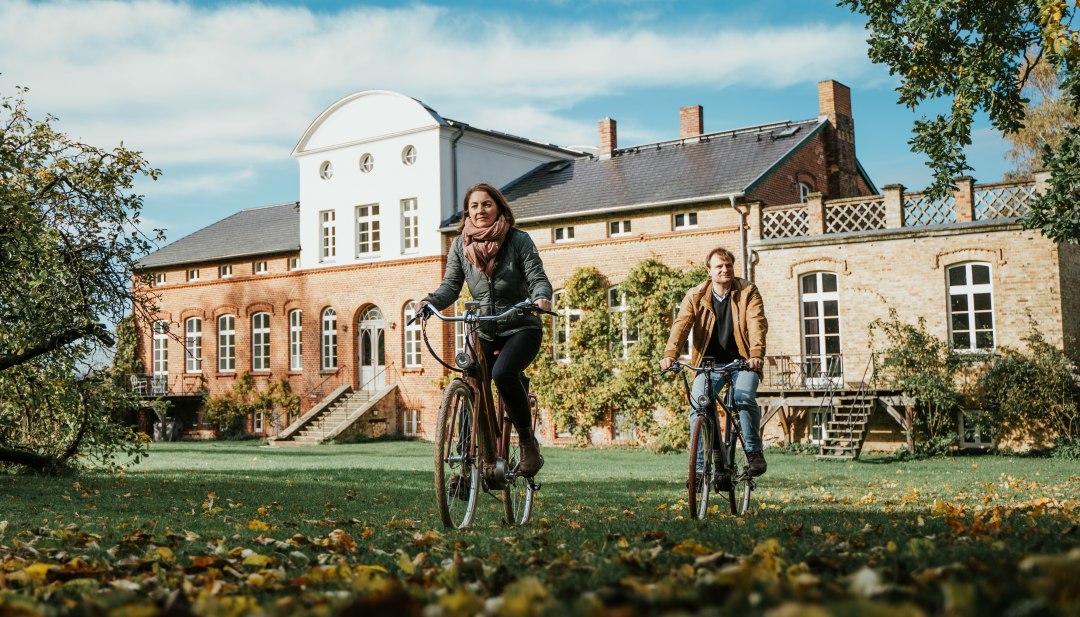 Dwie osoby jadą na rowerach wśród jesiennych liści przed zabytkowym dworem w Meklemburgii-Pomorzu Przednim.