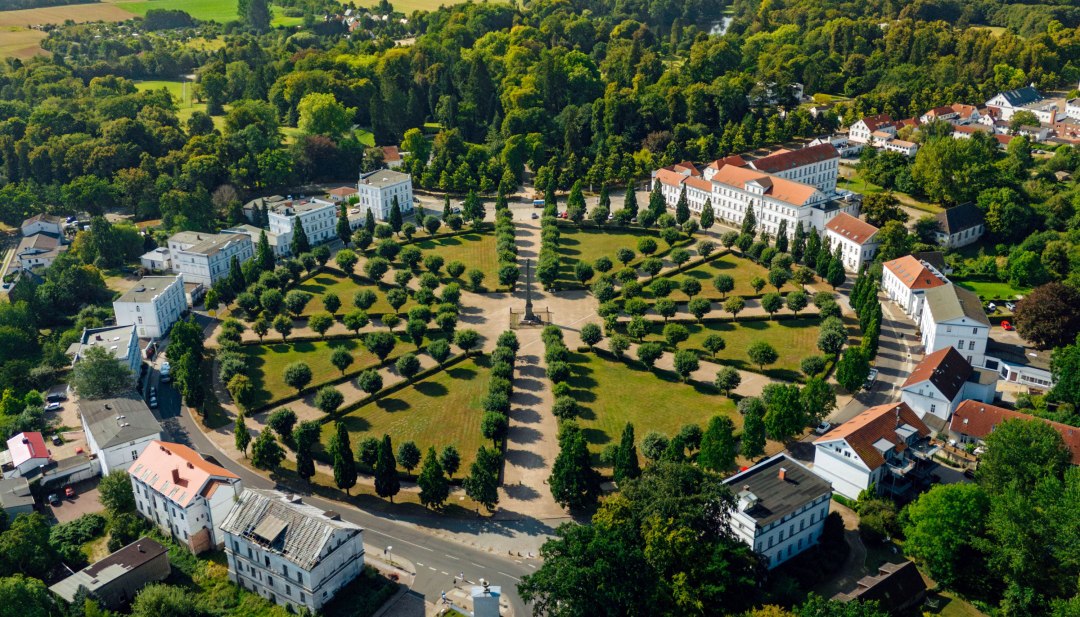 Luchtfoto van het Circus in Putbus op het eiland Rügen, een cirkelvormig plein met symmetrisch geplaatste bomen en classicistische gebouwen, omgeven door groene natuur en een landelijk landschap.