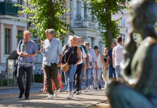 Grupa ludzi spaceruje brukowaną ścieżką z przewodnikiem miejskim. Po prawej stronie, niewyraźnie na zdjęciu, widać posąg nad stawem Pfaffenteich; w tle pojawiają się wille. W centrum uwagi znajduje się przewodnik miejski i para prowadząca ożywioną rozmowę., © Oliver Borchert
