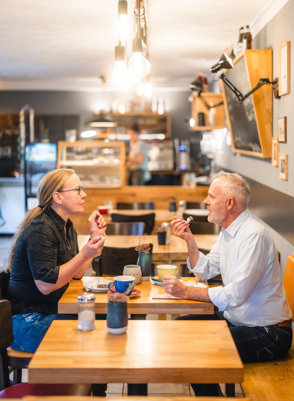 Twee mensen genieten van koffie en gebak in een gezellig caf&eacute; met moderne inrichting.