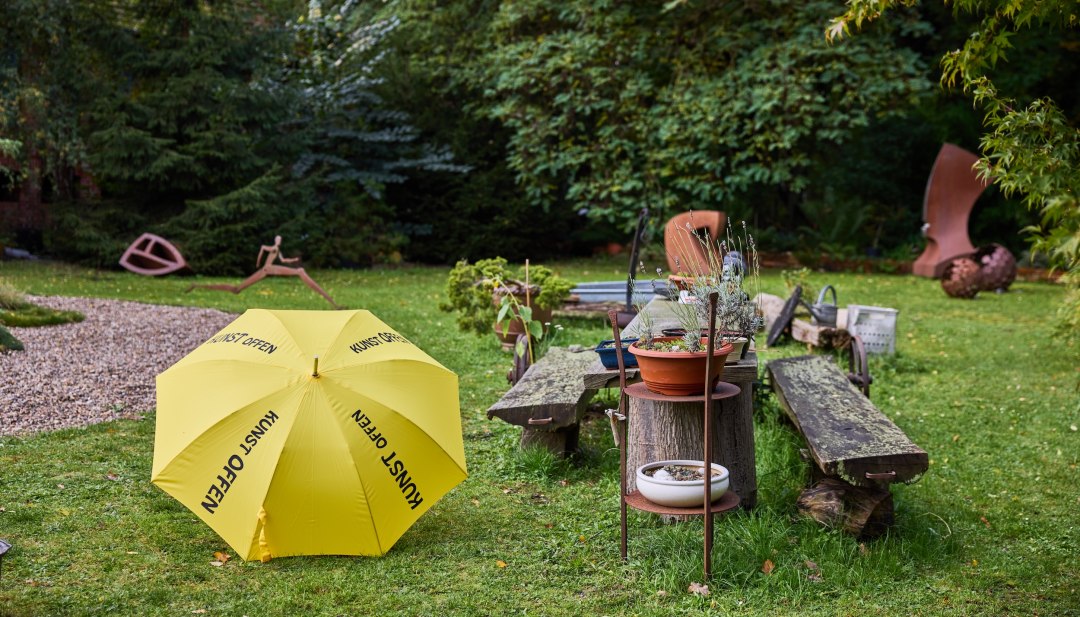Sculpture garden with yellow KunstOffen umbrella and metal sculptures at Kunst-Werk Kummer. In front of it is a yellow umbrella with the inscription "KunstOffen" // At Kunst-Werk Kummer in Kummer, Thomas Lehnigk opens his sculpture garden for KunstOffen. Between metal sculptures and lush green spaces, wooden benches invite you to linger. Art meets nature - a place that provides space for new perspectives with the typical feature - the yellow umbrella. // © TVMSN/Borchert Sculpture garden with yellow KunstOffen umbrella and metal sculptures at Kunst-Werk Kummer. In front of it is a yellow umbrella with the inscription "KunstOffen"