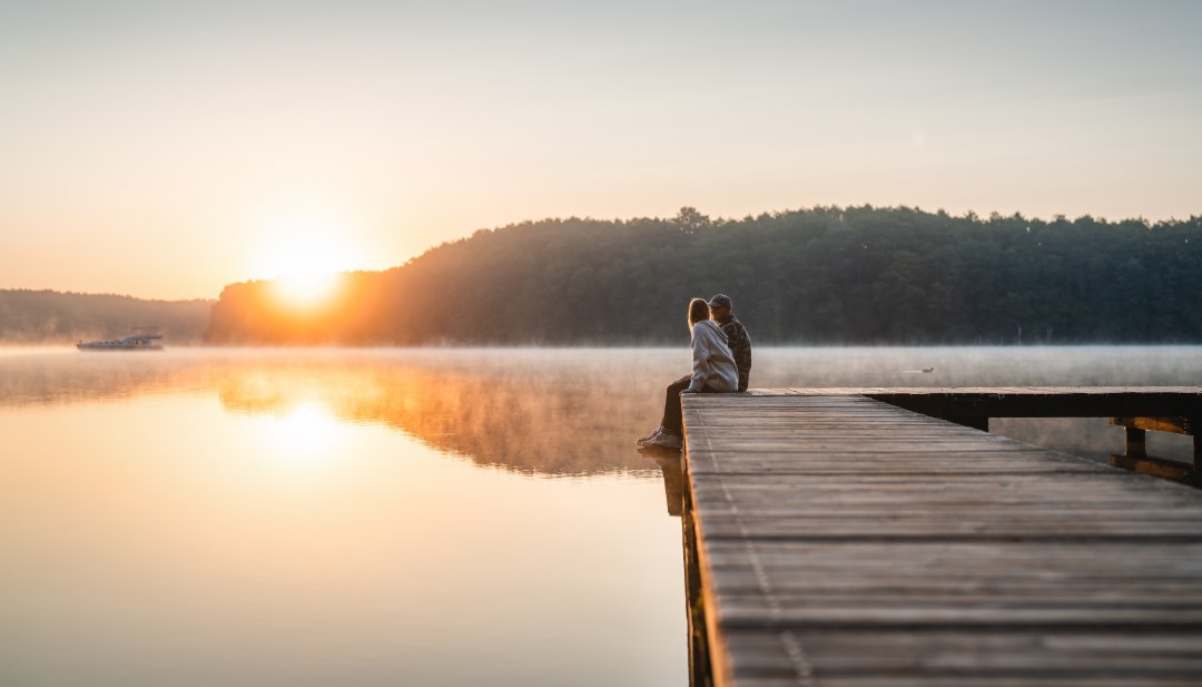 A couple sits on a wooden jetty by a calm lake and looks out at the sunrise, surrounded by morning mist.