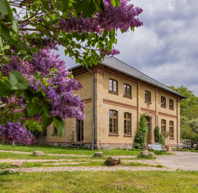 Lexow manor house with blooming lilacs in the foreground and historic brick façade. // Purple lilac blossoms frame the Lexow manor house, while the sky rises above the historic façade. The place breathes tranquillity amidst the greenery and flowers - a place to arrive and take a deep breath. // © Gutshaus Lexow Lexow manor house with blooming lilacs in the foreground and historic brick façade.