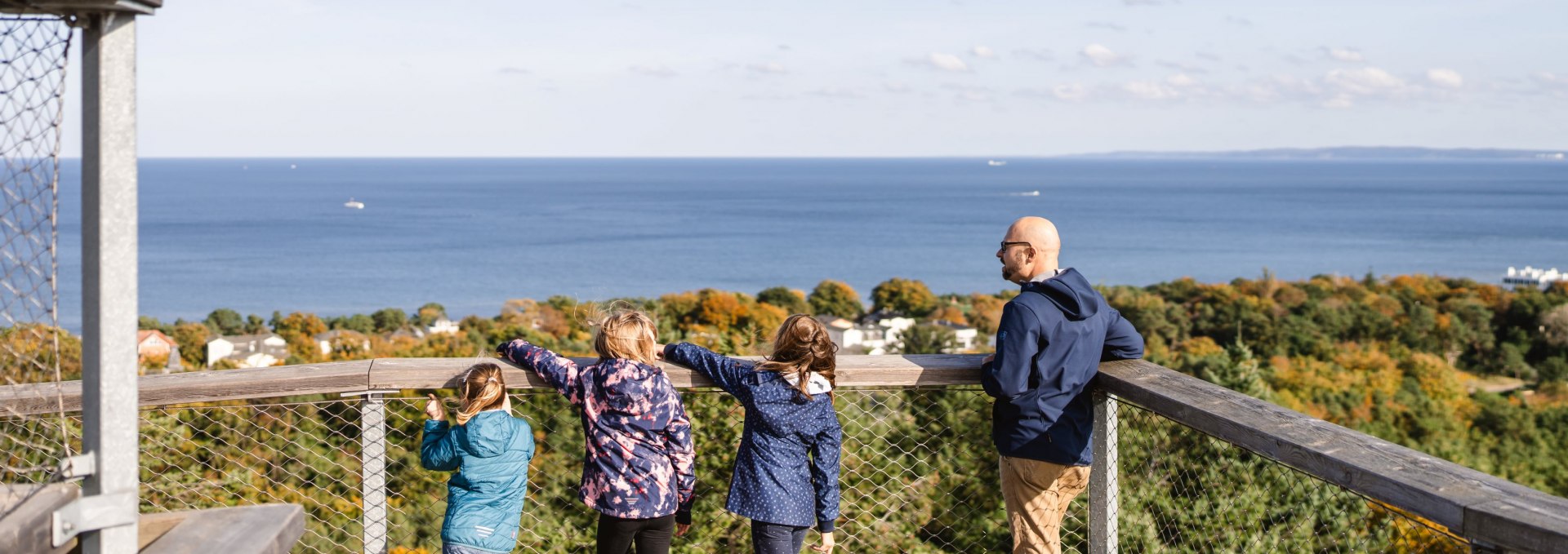  Twee volwassenen en drie kinderen staan op een uitkijkplatform en kijken uit over het bos naar de blauwe zee.