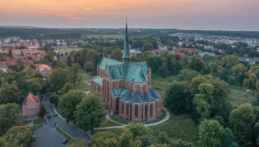Luchtfoto van de kathedraal in Bad Doberan bij zonsondergang, omringd door bomen, weiden en de omliggende stad.