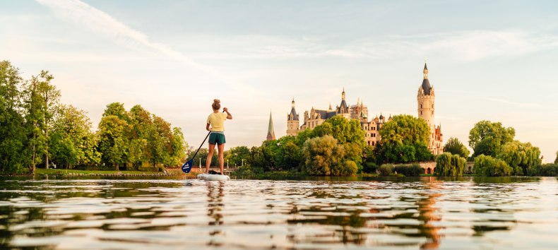 A person paddles on a lake in the warm evening light and looks out over a historic castle surrounded by lush greenery.