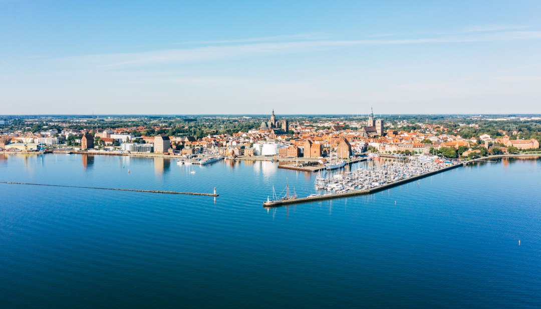 Luchtfoto van de Hanzestad Stralsund met haven, historische oude stad en Baltische Zee onder een blauwe hemel. // De Hanzestad Stralsund strekt zich uit tussen de haven en de historische oude stad. Baksteengotiek ontmoet zeilmasten, terwijl het water van de Oostzee helderblauw schittert. Hier kun je de maritieme geschiedenis en levendige havencultuur ontdekken - een plek die visie en aankomst combineert. // © MV-T/Gänsicke Luchtfoto van de Hanzestad Stralsund met haven, historische oude stad en Baltische Zee onder een blauwe hemel.