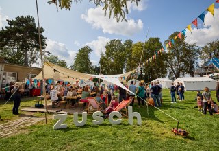 People stand under an awning decorated with colorful pennants // © Oliver Borchert People stand under an awning decorated with colorful pennants // © Oliver Borchert