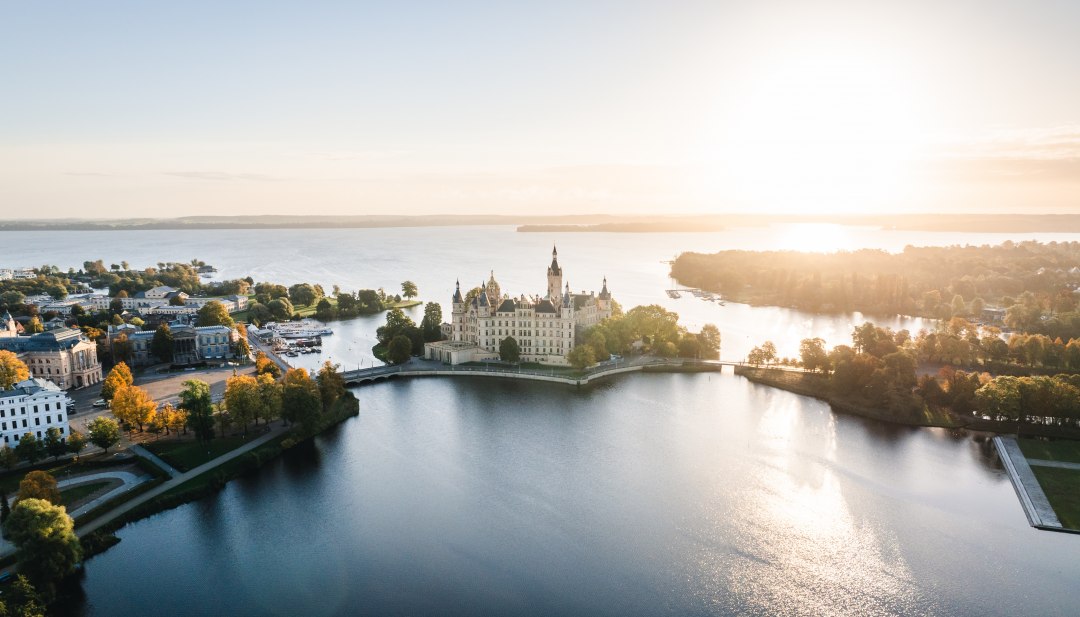 Kasteel Schwerin op een eiland in het meer bij zonsopgang, omringd door bomen en water. // De residentie op het kasteeleiland troont tussen water en lucht. Ochtendlicht raakt de torens van kasteel Schwerin, terwijl het silhouet wordt weerspiegeld in het kalme meer. Geschiedenis ontmoet uitgestrektheid - een plek die je ruimte geeft om te ademen en tegelijkertijd je blik gevangen houdt. // © MV-T/Gross Kasteel Schwerin op een eiland in het meer bij zonsopgang, omringd door bomen en water.