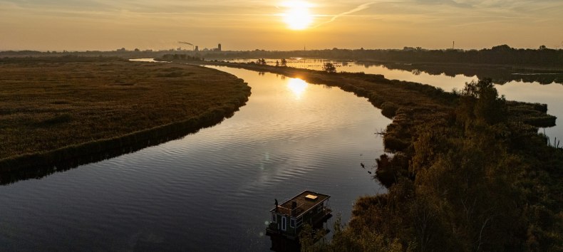 Woonboot op de Peene bij zonsondergang, omgeven door een vredig rivierlandschap en gouden licht aan de horizon.