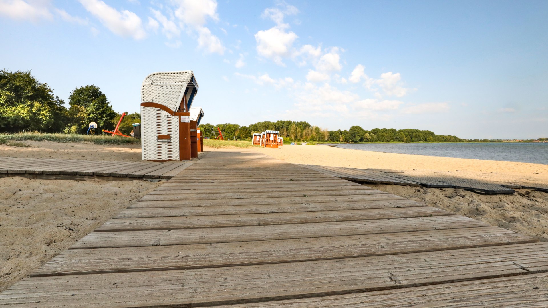 Drewniany chodnik prowadzi bez barier do leżak&oacute;w na piaszczystej plaży nad jeziorem Schaalsee, w otoczeniu spokojnej przyrody i błękitnego nieba.