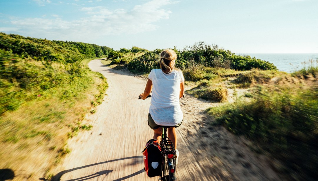 Vrouw fietst langs een kustpad door de duinen met uitzicht op de Baltische Zee in de zon.