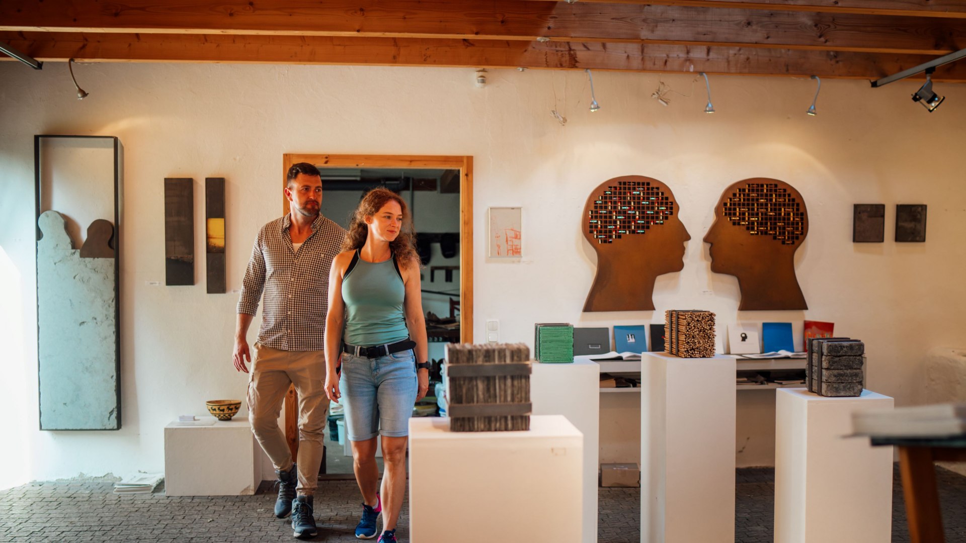 Een koppel wandelt door een kunsttentoonstelling in de oude watermolen in Wreechen. De tentoonstelling bestaat uit sculpturen en kunstwerken, waaronder twee silhouetten van menselijke hoofden met gedetailleerde ontwerpen. De warme verlichting en rustieke sfeer scheppen een uitnodigende sfeer.