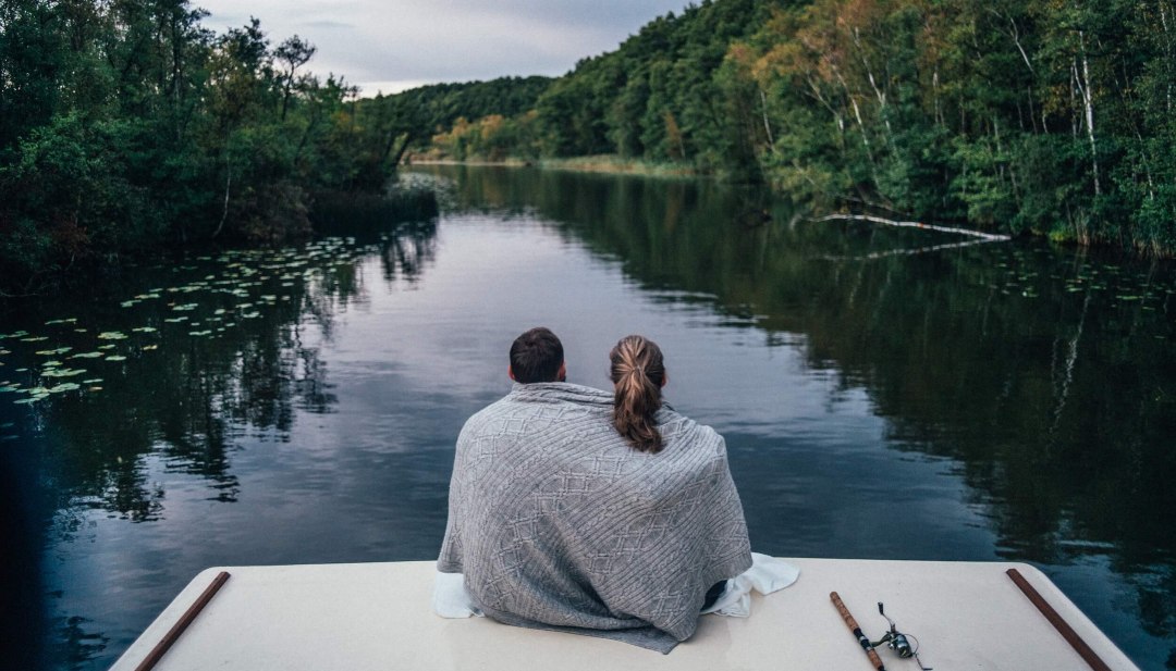A couple sits on a houseboat, wrapped in a blanket, looking out over a quiet, wooded river in the Mecklenburg Lake District. // Peace for two - cuddled up in the silence on a houseboat in the idyllic nature of the Mecklenburg Lake District. // © MV-T/Gänsicke A couple sits on a houseboat, wrapped in a blanket, looking out over a quiet, wooded river in the Mecklenburg Lake District.