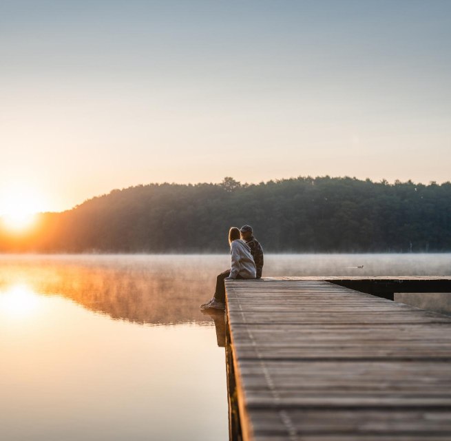A couple sits on a jetty at sunset on Lake Mirov. A houseboat passes by in the background.