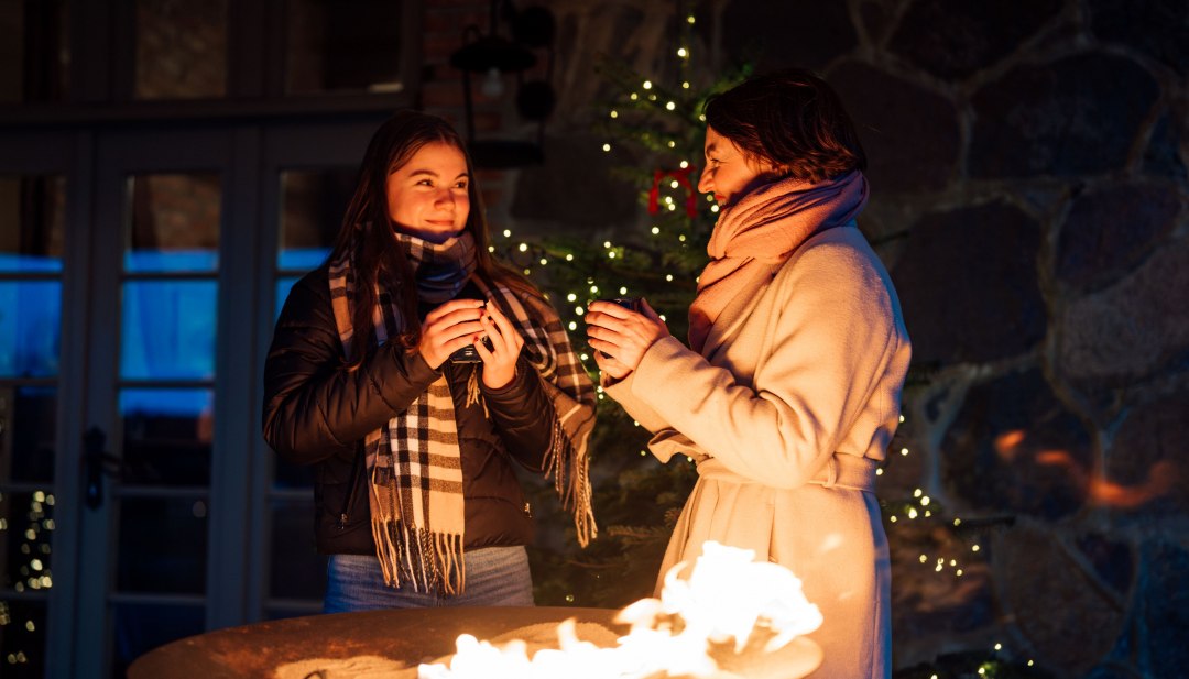 Two women warm themselves by a fire bowl at the Advent market in Ulrichshusen Castle, surrounded by twinkling lights and a wintry atmosphere.