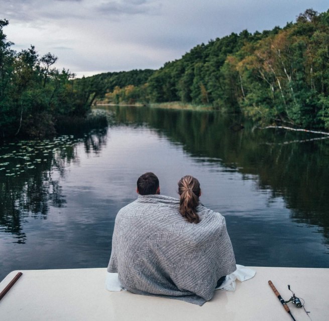 A couple sits on a houseboat, wrapped in a blanket, looking out over a quiet, wooded river in the Mecklenburg Lake District.