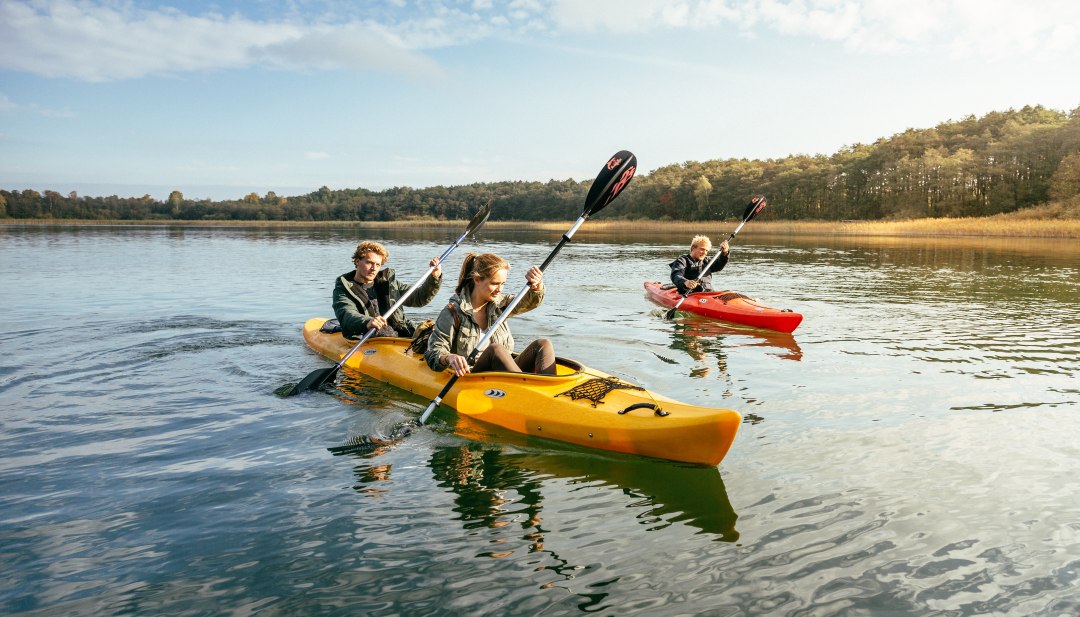 Peddelavonturen in een natuurparadijs - geniet tijdens het kajakken van de rust van het merenlandschap en ontdek de waterwegen van Mecklenburg-Vorpommern., © TMV/Roth Drie jonge mensen peddelen met hun kajak over een rustig meer midden op het platteland van Mecklenburg-Vorpommern.