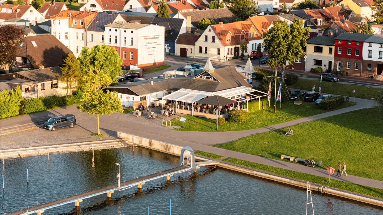 Restauracja DE ZEES w Ribnitz-Damgarten widziana z lotu ptaka – taras nad brzegiem, ścieżki prowadzące do lokalu i zachęcające do odpoczynku. // © Ribnitzer Fischhafen/Hugo Scholz-Witzel Restauracja DE ZEES w Ribnitz-Damgarten widziana z lotu ptaka – taras nad brzegiem, ścieżki prowadzące do lokalu i zachęcające do odpoczynku. // © Ribnitzer Fischhafen/Hugo Scholz-Witzel