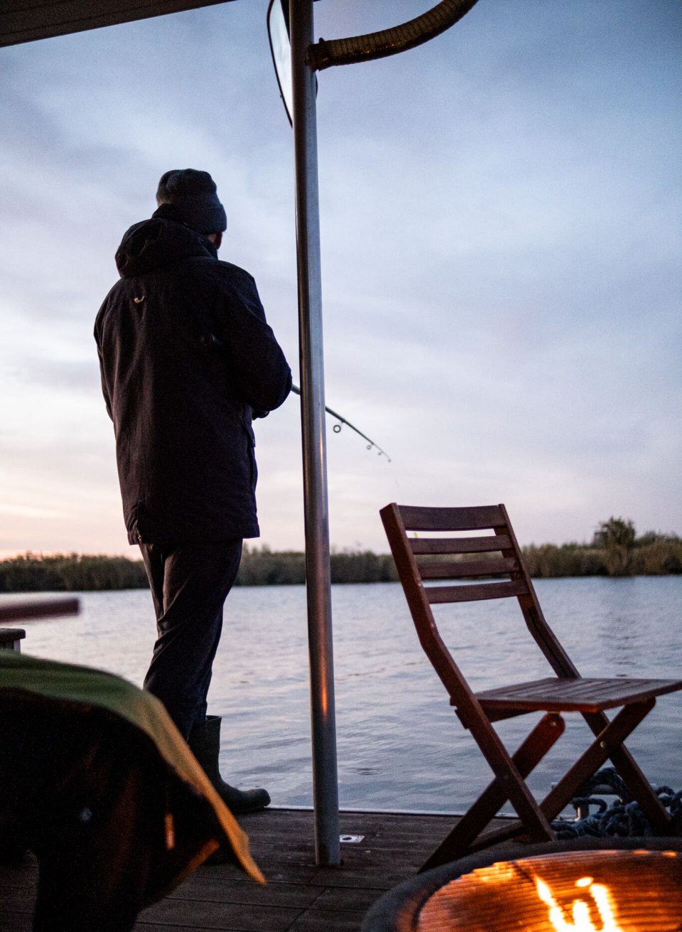 Persoon aan het vissen vanaf het dek van een woonboot op de Peene bij zonsondergang, met een open haard op de voorgrond.