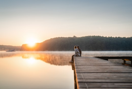 A couple sits on a wooden jetty by a calm lake and looks out at the sunrise, surrounded by morning mist.