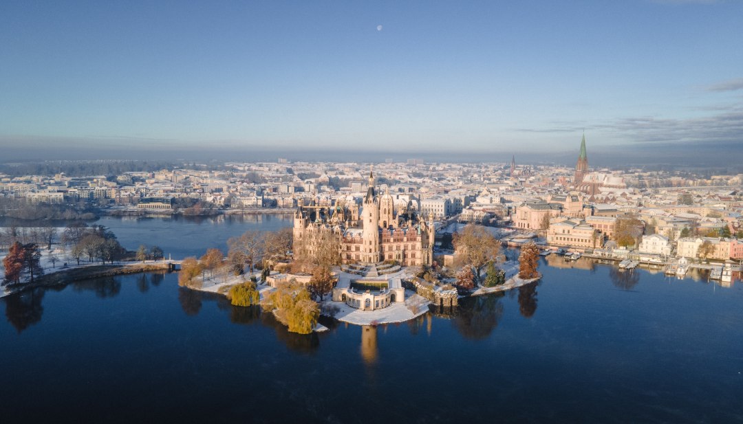 Winters sprookjeskasteel - Kasteel Schwerin op zijn eiland schittert te midden van besneeuwde landschappen en weerspiegelend water als herkenningspunt van de deelstaathoofdstad Mecklenburg-Vorpommern., © TMV/Scholz-Witzel Luchtfoto van het besneeuwde kasteel van Schwerin op een eiland in het meer met uitzicht op de oude stad in de winter.
