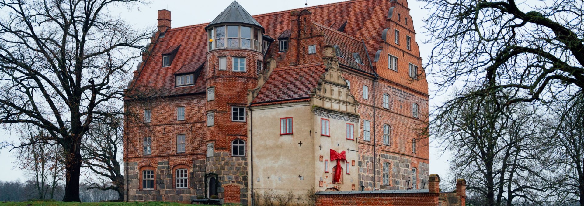 Buitenaanzicht van het kasteel Ulrichshusen in Mecklenburg, versierd voor Kerstmis met een rood lint, omringd door bomen en een groene winterweide.