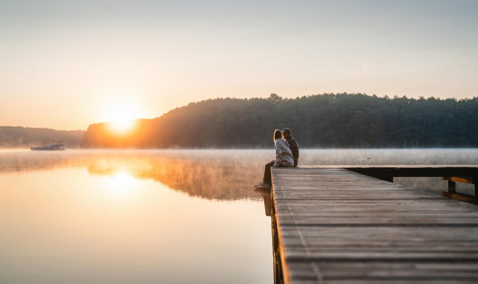 A couple sits on a jetty at sunset on Lake Mirov. A houseboat passes by in the background. // Sitting on the jetty enjoying the evening on Lake Mirower See // © MV-T/Gross A couple sits on a jetty at sunset on Lake Mirov. A houseboat passes by in the background.