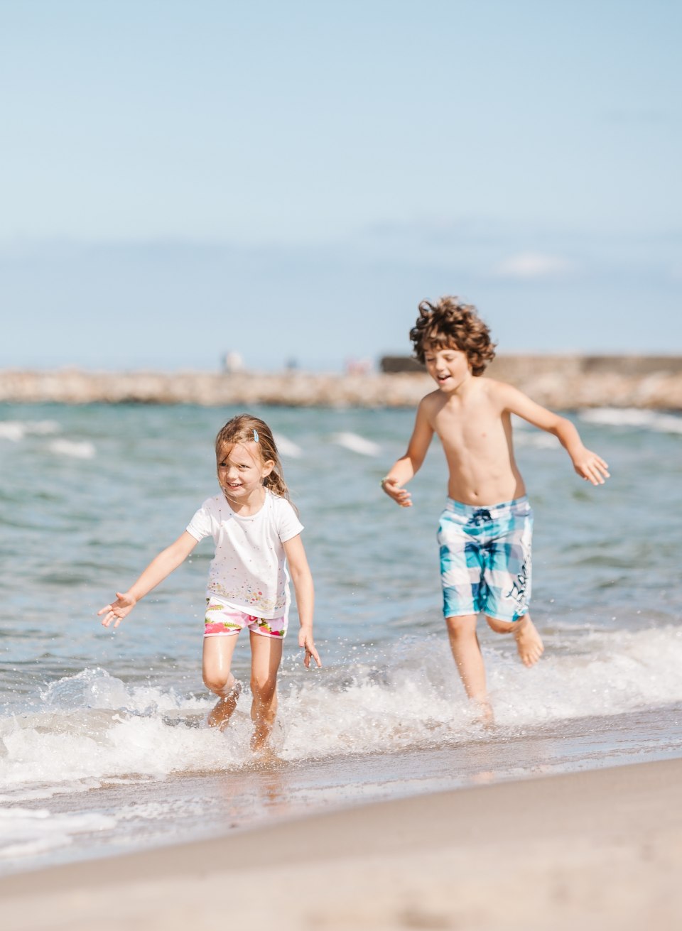 Dwoje dzieci biegnie śmiejąc się przez płytkie bałtyckie fale na plaży, z zieloną latarnią morską na molo w tle.