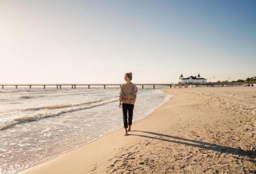 Kobieta spacerująca po plaży Ahlbeck o wschodzie słońca z molo w tle // © MV-T/Petermann Kobieta spacerująca po plaży Ahlbeck o wschodzie słońca z molo w tle