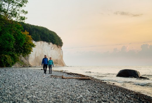 Para spaceruje wzdłuż kamienistej plaży z widokiem na morze o wschodzie słońca na kredowym wybrzeżu Rugii. // Świeże bałtyckie powietrze i malownicze widoki - para cieszy się ciszą i spokojem podczas porannego spaceru wzdłuż kredowego wybrzeża na wyspie Rugia. Idealne miejsce dla miłośników przyrody i pieszych wędrówek. // © MV-T/Petermann Para spaceruje wzdłuż kamienistej plaży z widokiem na morze o wschodzie słońca na kredowym wybrzeżu Rugii.
