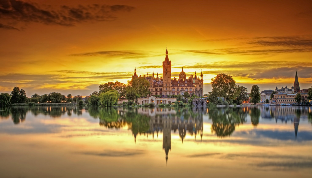 Kasteel Schwerin wordt omlijst door de laatste zonnestralen. De zonsondergang kleurt de lucht oranje. Kasteel Schwerin wordt weerspiegeld in het kalme meer.