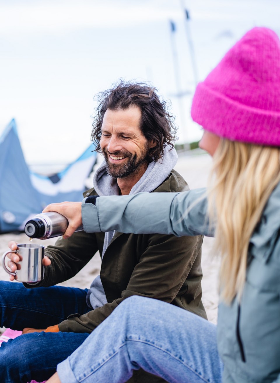 A couple sits on the beach in Timmendorf, smiling and enjoying a hot cup of coffee with a kite in the background.