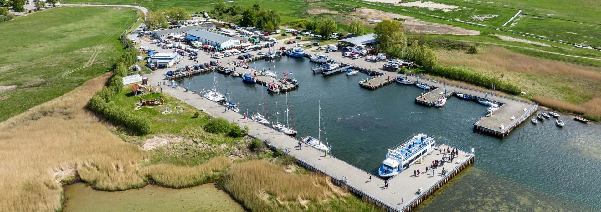 Luchtfoto van de haven in Thiessow met aanlegplaatsen voor boten en schepen, groene weiden op de achtergrond en de Baltische Zee onder een heldere hemel.