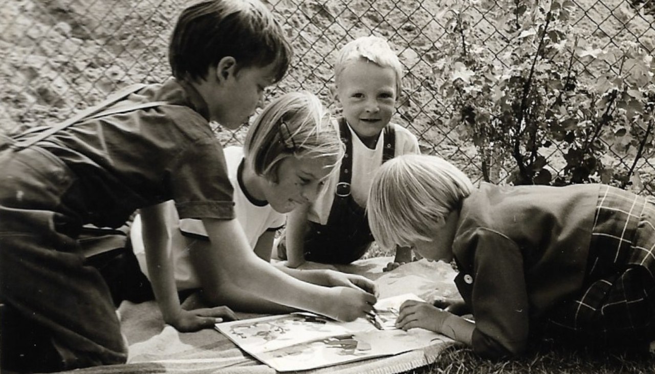Czarno-białe zdjęcie czwórki dzieci leżących na kocu na łące przed płotem i malujących obrazek., © privat
