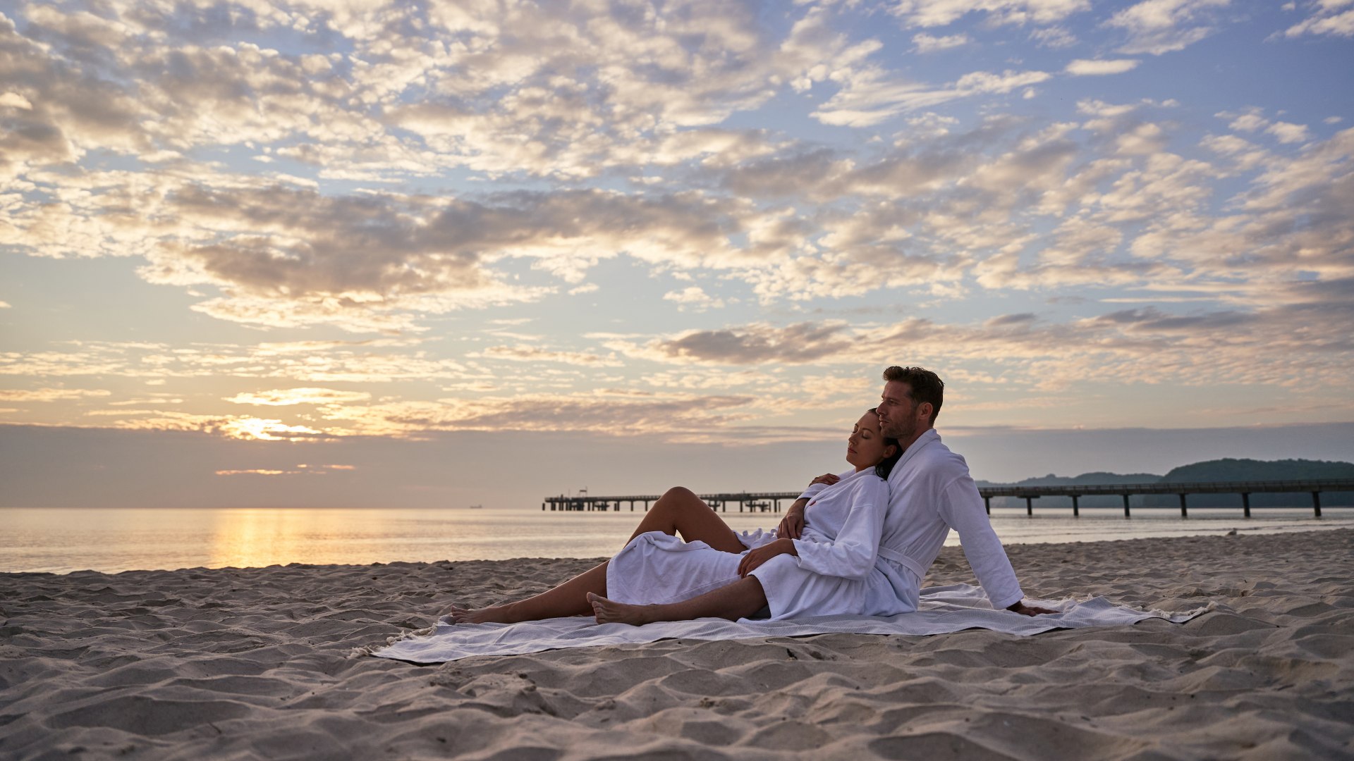 Two people sitting on the beach in bathrobes with the Binz pier in the background