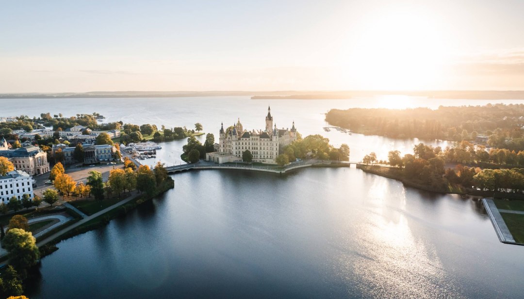 Widok z lotu ptaka na zamek Schwerin na wyspie na jeziorze, otoczony wodą i drzewami o wschodzie słońca. // Romantyczna atmosfera nad jeziorem Schwerin - bajkowy zamek Schwerin lśni w porannym świetle i zaprasza do spędzenia niezapomnianych chwil w Meklemburgii-Pomorzu Przednim jesienią. // © MV-T/Gross Widok z lotu ptaka na zamek Schwerin na wyspie na jeziorze, otoczony wodą i drzewami o wschodzie słońca.