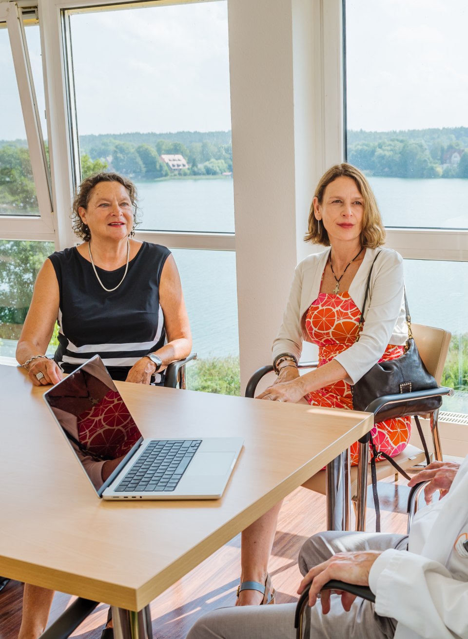Twee vrouwen zitten aan een tafel in een kamer en hebben een gesprek met een arts.