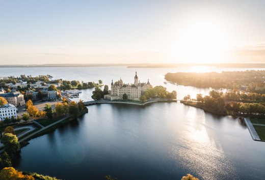 Schwerin Castle on an island in the lake at sunrise, surrounded by trees and water.